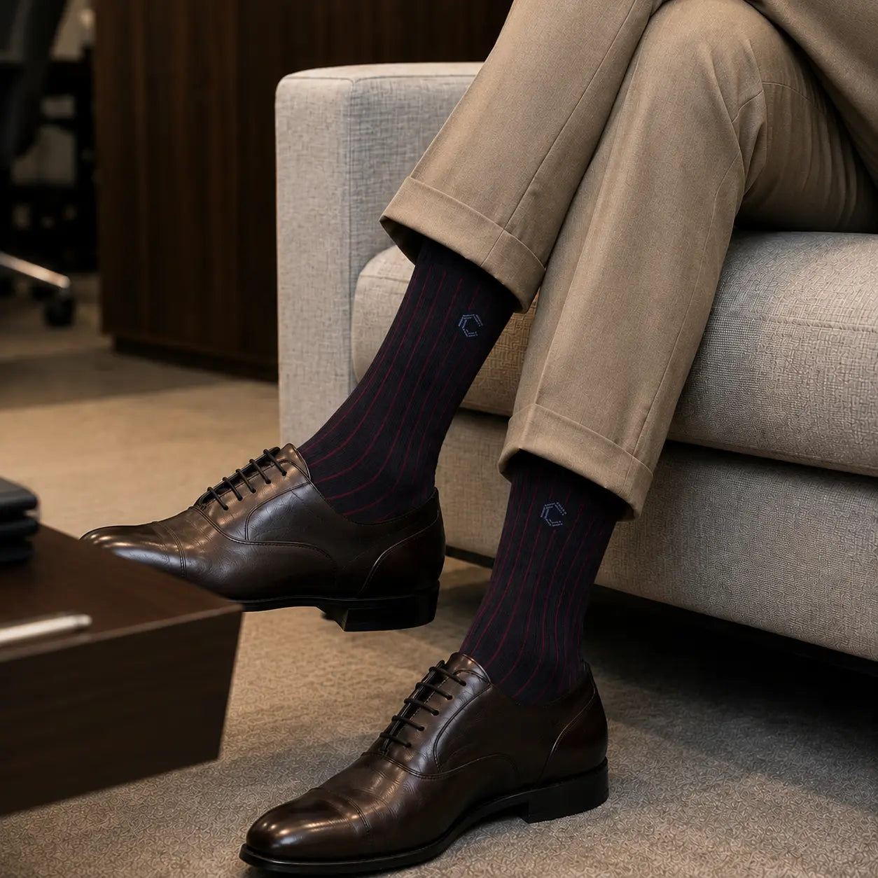 A person seated on a light gray sofa wearing dark brown oxford shoes, beige trousers, and dark navy striped socks with a small embroidered logo.