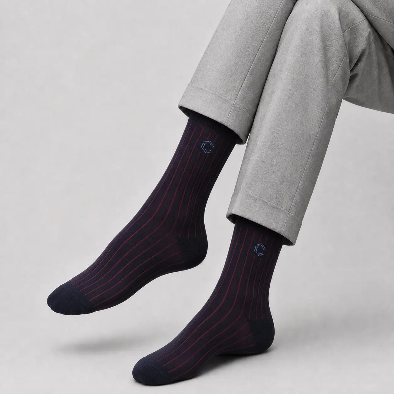 A pair of dark navy crew socks with thin red pinstripes, worn on a person's feet with light gray trousers, shown against a neutral light gray background.